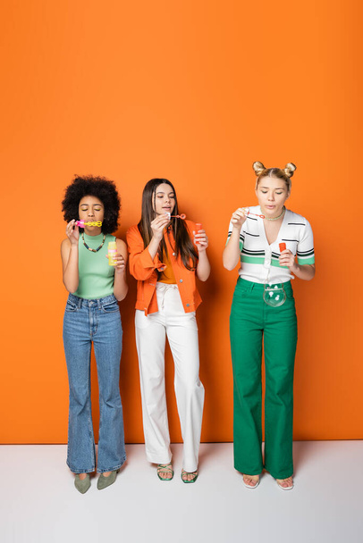 Full length of trendy and multiethnic teenage girlfriends with colorful makeup wearing casual outfits while blowing soap bubbles and standing on orange background, trendy and stylish hairstyles - Photo, Image