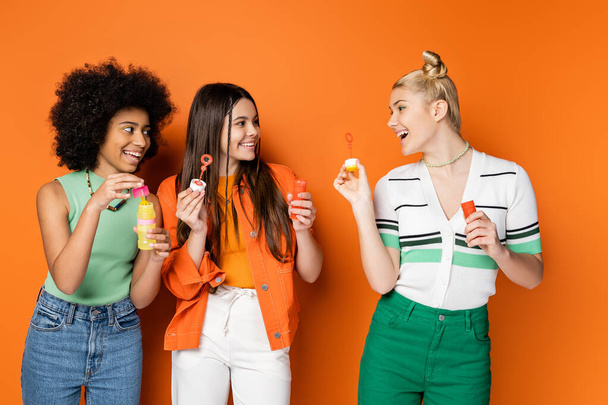 Positive blonde teenager with bold makeup holding soap bubbles and talking to multiethnic girlfriends in casual clothes while standing on orange background, trendy and stylish hairstyles - Photo, Image