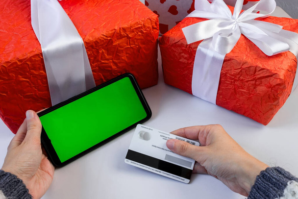 Female hands in a sweater hold a smartphone with a green horizontal screen and a payment card near the gift boxes on the table. The concept of holiday shopping on the Internet . Chroma key. - Фото, зображення