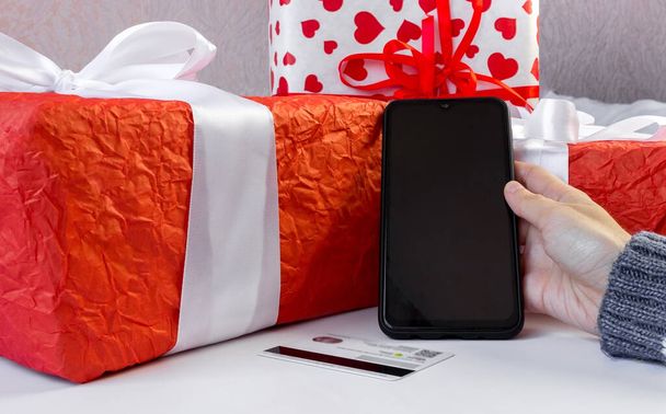 Woman's hands in a sweater hold a smartphone and a payment card near the gift boxes on the table. Online payment. Commerce and internet banking concept. - Photo, Image