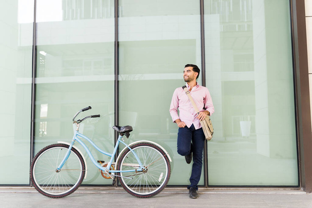 Χαρούμενος χαλαρός νεαρός που απολαμβάνει την πόλη μετά το ταξίδι του για να εργαστεί με το φιλικό προς το περιβάλλον ποδήλατό του ακουμπώντας στο αστικό κτίριο - Φωτογραφία, εικόνα