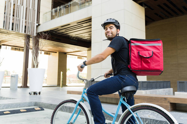 Giovane latino che sorride e va in bicicletta in città mentre lavora per un servizio di consegna di cibo dall'aspetto felice - Foto, immagini