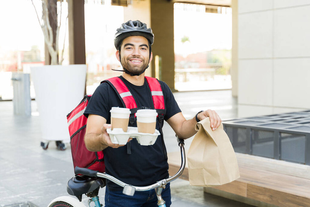 Excited attractive man delivering coffee and food lunch while smiling and working in the delivery service riding a bicycle - Фото, изображение