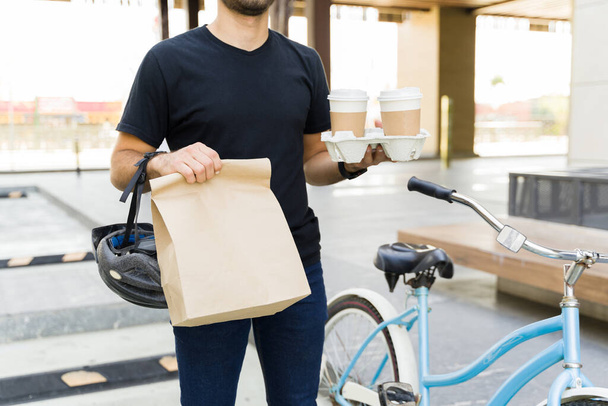Close up of a young man carrying coffee and a lunch bag while working for a delivery app delivering food using a bike in the city - Фото, изображение