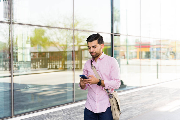 Aantrekkelijke Latijnse man loopt in de stad op zijn woon-werkverkeer naar zijn werk gebouw tijdens het sms 'en op zijn smartphone  - Foto, afbeelding