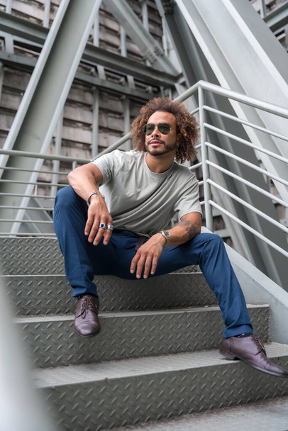 Young man with afro hair wearing sunglasses on the stairs in the city sitting looking to the left, urban fashion cool concept of a hipster guy with stylish - Foto, Imagen