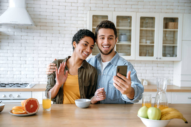 Cheerful loving couple of different nationalities, talking via video conference using smart phone, during a morning breakfast, looking into the camera of the mobile phone, smiling happily - Fotografie, Obrázek
