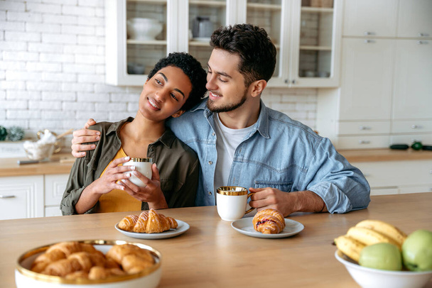 Happy spouses of different nationalities, sit und hugging at home in the kitchen, dressed in stylish clothes, drinking morning coffee with croissants, dreaming about something, look away, smile - Foto, Imagem