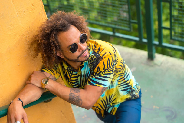 Past of an attractive man with afro hair in a summer summer palm tree shirt on a yellow background and a ladder - Фото, зображення