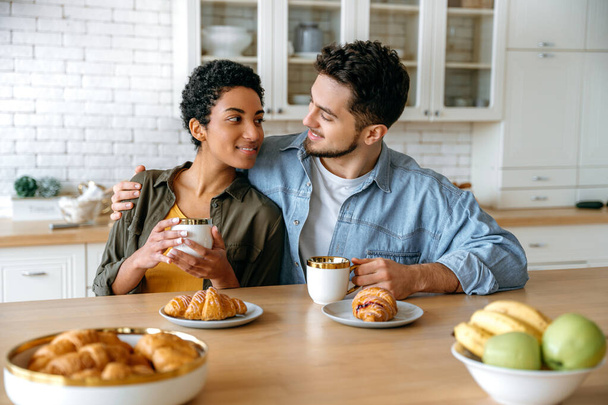 Pozytywna para zakochana w różnych narodowościach, siedząca i przytulająca się w domu w kuchni, ubrana w stylowe ubrania, pijąca poranną kawę z rogalikami, patrząca na siebie, uśmiechająca się - Zdjęcie, obraz