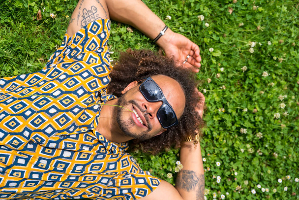 Afro hair man smiling with sunglasses in summer enjoying a holiday or vacation day lying on the grass - Foto, imagen
