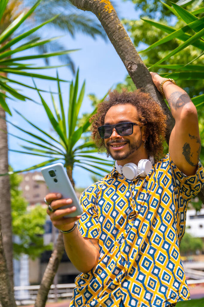 Πορτρέτο του αφρο-μαλλιά άνθρωπος στις καλοκαιρινές διακοπές δίπλα σε κάποια φοίνικες από την παραλία κοιτάζοντας το κινητό. Έννοια ταξιδιών και τουρισμού - Φωτογραφία, εικόνα