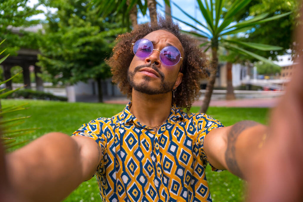 Afro-haired man on summer vacation next to some palm trees next to the beach taking a selfie with both hands. Travel and tourism concept - Photo, Image