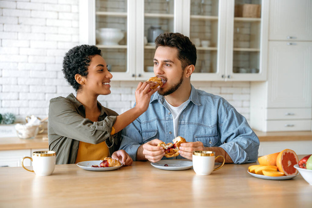 Joyful couple in love of different nationalities, happy spouses, sit at home in the kitchen, drinking morning coffee with croissants, woman feeding a man a croissant, look at each other, smile - Photo, Image