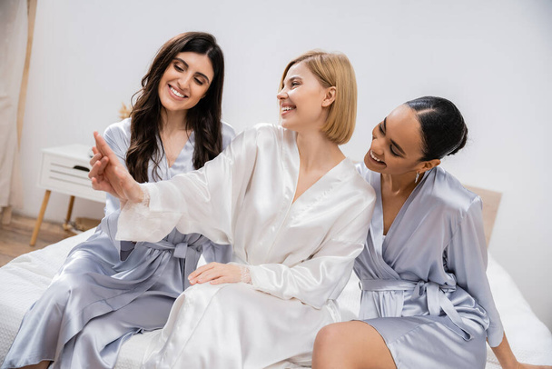 happy blonde bride showing engagement ring to her multicultural bridesmaids, sitting on bed together, bridal shower, silk robes, brunette and blonde women, diversity, multicultural best friends  - Photo, Image