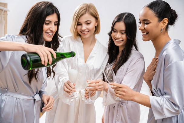 gelukkig vrouw gieten champagne in glazen in de buurt van interraciale vriendinnen, feest, vrolijke bruid en bruidsmeisjes, brunette en blond, diversiteit, bruids douche, beste vrienden, vier vrouwen  - Foto, afbeelding