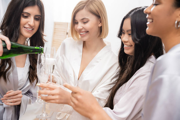 happy woman pouring champagne into glasses near multicultural girlfriends, celebration, cheerful bride and bridesmaids, brunette and blonde, diversity, bridal shower, best friends, four women  - Foto, Imagem