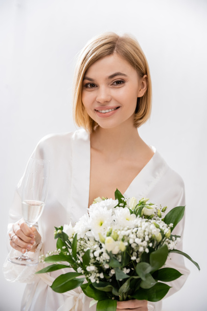 fröhliche Braut mit blonden Haaren im weißen Seidenmantel lächelnd, mit einem Glas Champagner und Brautstrauß in der Hand, junge Frau, schön, aufregend, feminin, selig, Porträt  - Foto, Bild