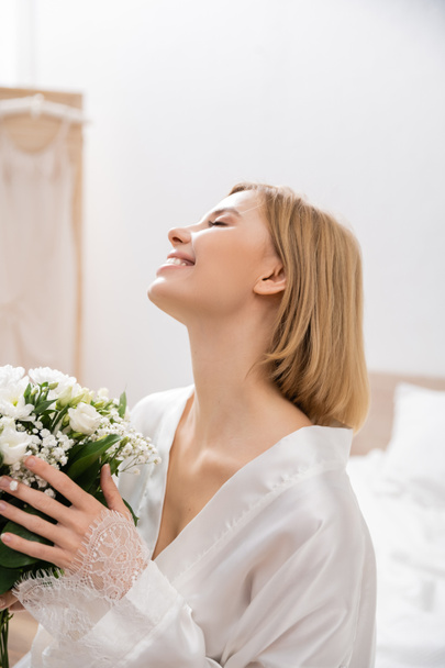 happiness, cheerful bride with blonde hair sitting on bed and holding bridal bouquet, young woman in white robe, beautiful, excitement, feminine, blissful, wedding preparation, white flowers  - Photo, Image