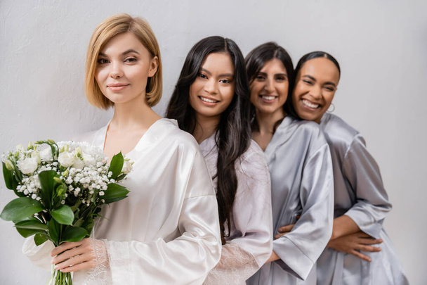 positivity, bride and bridesmaids, happy woman holding bouquet of flowers and standing near cheerful interracial friends on grey background, racial diversity, silk robes, fashion  - Photo, Image