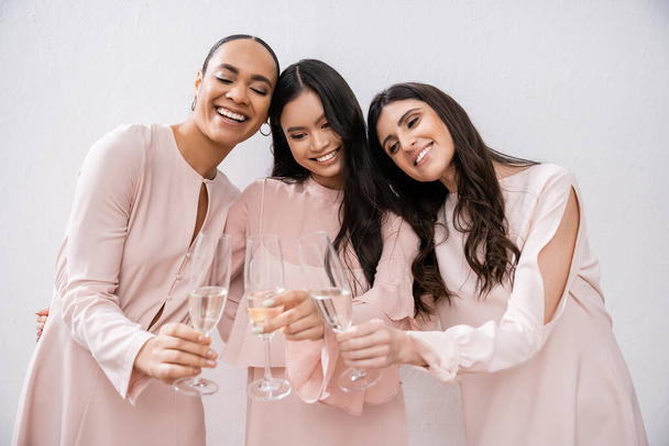 tres damas de honor multiculturales, mujeres bonitas en vestidos de color rosa pastel tintineando copas de champán sobre fondo gris, diversidad cultural, moda, celebración, hurras  - Foto, imagen