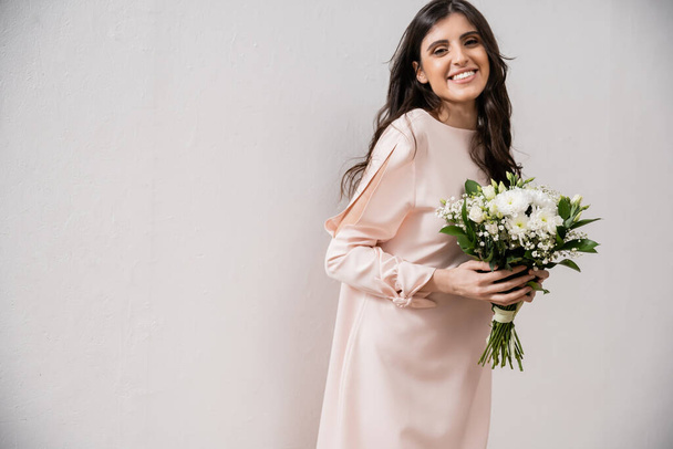 cheerful bridesmaid in pastel pink dress holding bridal bouquet, brunette woman on grey background, white flowers, special occasion, wedding, fashion, smile and joy, looking at camera  - Foto, imagen