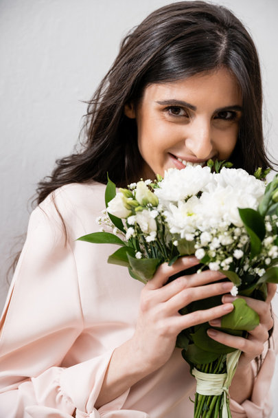 speciální příležitost, šťastná družička v pastelové růžové šaty drží kytice, brunetka žena na šedém pozadí, bílé květy, svatba, móda, úsměv a radost, při pohledu do kamery  - Fotografie, Obrázek