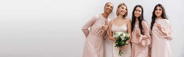 積極的で幸せな花嫁のウェディングドレスはブライダルブーケを保持し、グレーの背景に異人種間の花嫁介添人の近くに立って、シャンパングラス、人種の多様性、ファッション、バナー  - 写真・画像
