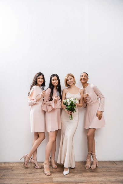 positivity, blonde bride in wedding dress holding bouquet and standing near interracial bridesmaids with champagne glasses on grey background, racial diversity, fashion, multicultural young women  - Photo, Image