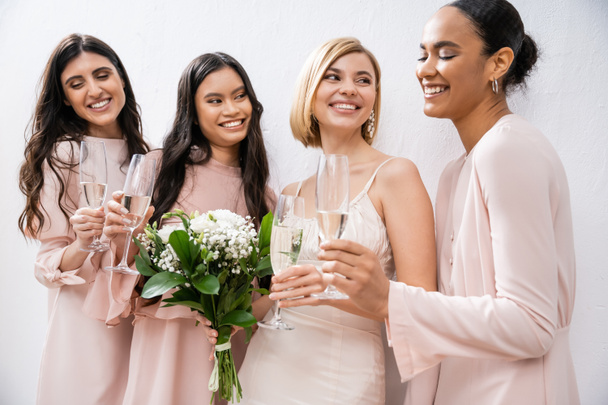 позитивность, блондинка невеста в свадебном платье держа букет, стоя с межрасовыми подружками невесты, бокалы шампанского, серый фон, расовое разнообразие, мода, мультикультурные молодые женщины  - Фото, изображение