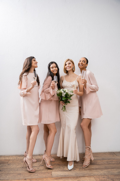 joy, blonde bride in wedding dress holding bouquet, standing with interracial bridesmaids, champagne glasses, grey background, racial diversity, fashion, multicultural young women  - Foto, Imagem