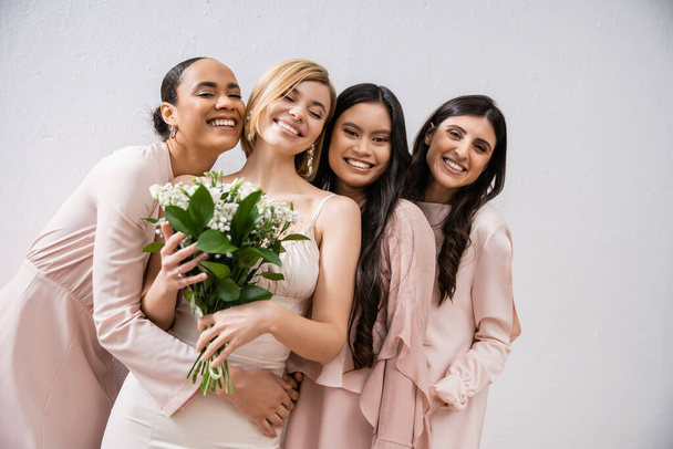 позитивность, жизнерадостные межрасовые подружки невесты, обнимающие счастливую невесту в свадебном платье, свадебный букет, серый фон, расовое разнообразие, мода, брюнетки и блондинки, белые цветы  - Фото, изображение