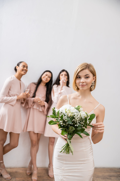beautiful bride in wedding dress holding bridal bouquet, standing near blurred interracial bridesmaids on grey background, happiness, special occasion, blonde and brunette women  - Photo, Image