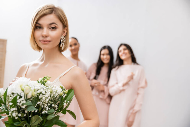 attractive bride in wedding dress holding bridal bouquet, standing near blurred interracial bridesmaids on grey background, happiness, special occasion, blonde and brunette women  - Photo, Image