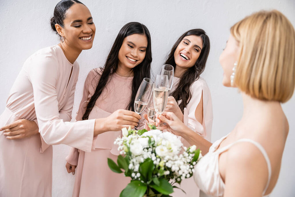 mulheres multiculturais felizes copos clinking com champanhe, noiva com flores brancas, mulheres morenas e loiras, damas de honra, diversidade, positividade, buquê de noiva, fundo cinza  - Foto, Imagem