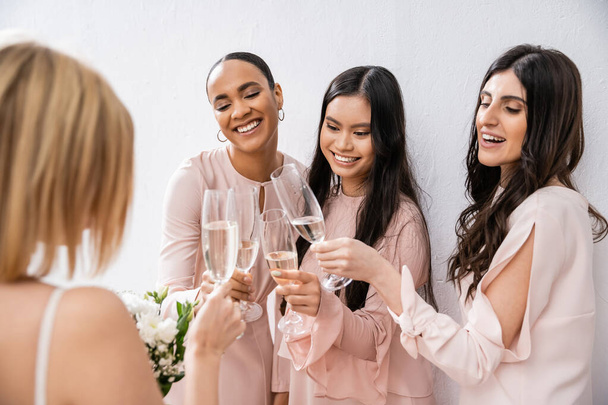 vrolijke multiculturele vriendinnen klinkende glazen met champagne, bruid met witte bloemen, brunette en blonde vrouwen, bruidsmeisjes, diversiteit, positiviteit, bruidsboeket, grijze achtergrond  - Foto, afbeelding