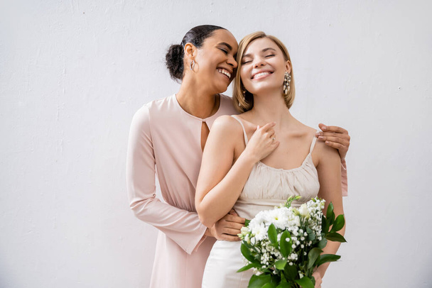 special occasion, cheerful bride with bridesmaid, happy interracial women, wedding dress and bridesmaid gown, african american woman hugging engaged friend on grey background  - Fotó, kép