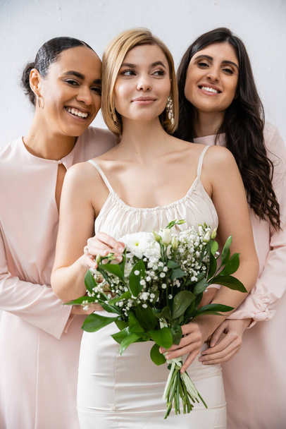 special occasion, brunette and interracial bridesmaids hugging blonde bride, friendship goals, grey background, happy multicultural girlfriends, cultural diversity, bridal bouquet, bridal party  - Photo, Image