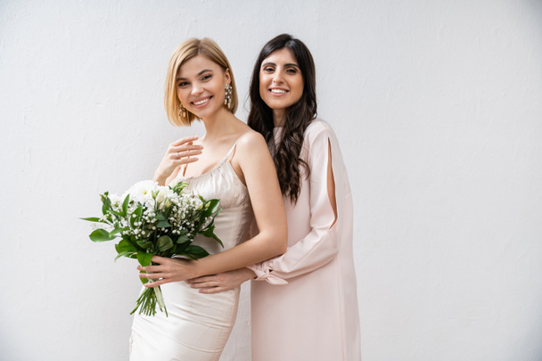 special occasion, cheerful bridesmaid hugging bride, friendship goals, grey background, happy girlfriends, bridal bouquet, blonde and brunette women, white flowers, happiness  - Photo, Image