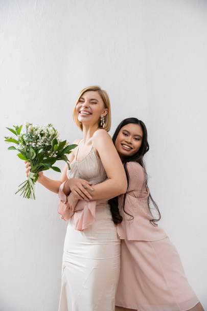 special occasion, asian bridesmaid hugging beautiful bride, friendship goals, grey background, happy girlfriends, bridal bouquet, blonde and brunette women, white flowers, happiness  - Photo, Image