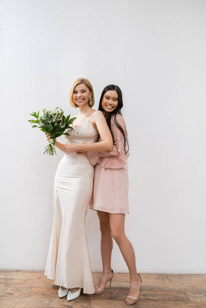 special occasion, cheerful asian bridesmaid hugging beautiful bride, friendship goals, grey background, happy girlfriends, bridal bouquet, blonde and brunette women, white flowers, happiness  - Photo, Image