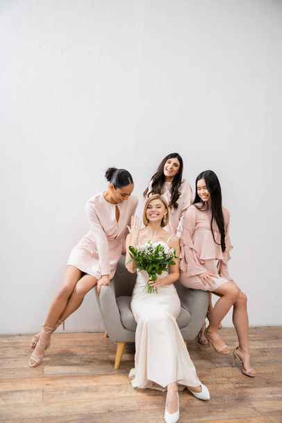 wedding photography, cultural diversity, four women, joyful bride with bouquet showing her engagement ring near bridesmaids, wedding day, sitting on armchair, grey background, happiness and joy  - Photo, Image