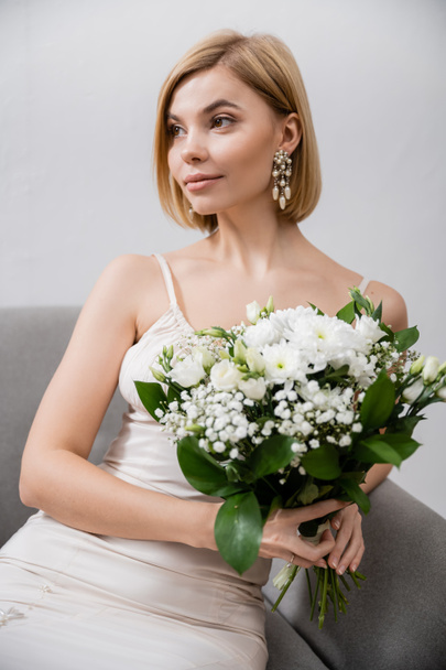 special occasion, beautiful and blonde bride in wedding dress sitting in armchair and holding bouquet on grey background, white flowers, bridal accessories, feminine, blissful  - Фото, изображение