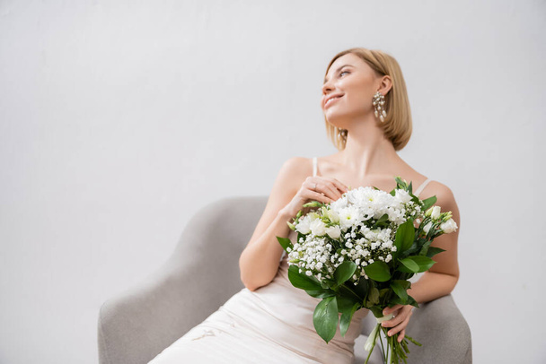 special occasion, happy blonde bride in wedding dress sitting in armchair and holding bouquet on grey background, engagement ring, white flowers, bridal accessories, happiness, feminine  - Foto, immagini