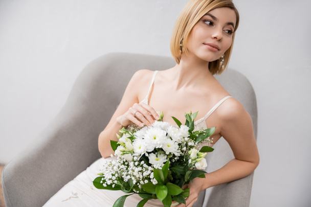 特別な機会に、アームチェアに座って灰色の背景、婚約指輪、白い花、ブライダルアクセサリー、幸福、女性の花束を保持するウェディングドレスの美しい若い花嫁  - 写真・画像