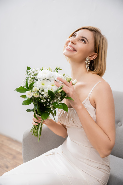 attractive and blonde bride in wedding dress sitting in armchair and holding bouquet on grey background, white flowers, bridal accessories, happiness, special occasion, smiling, feminine, blissful  - Photo, Image