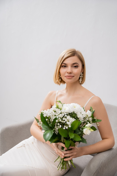 κομψή και ξανθιά νύφη με νυφικό που κάθεται στην πολυθρόνα και κρατά μπουκέτο σε γκρι φόντο, λευκά λουλούδια, νυφικά αξεσουάρ, ευτυχία, ειδική περίσταση, όμορφη, θηλυκή, μακάρια  - Φωτογραφία, εικόνα