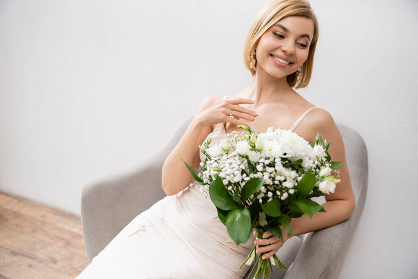 cheerful and elegant bride in wedding dress sitting in armchair and holding bouquet on grey background, white flowers, bridal accessories, happiness, special occasion, beautiful, feminine, blissful  - Foto, afbeelding