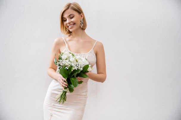 χαρούμενη και ξανθιά νύφη με νυφικό που κρατά μπουκέτο σε γκρι φόντο, λευκά λουλούδια, νυφικά αξεσουάρ, ευτυχία, ειδική περίσταση, όμορφη, θηλυκή, μακάρια  - Φωτογραφία, εικόνα