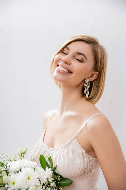 beautiful and blonde bride in wedding dress holding bouquet on grey background, joy, white flowers, bridal accessories, happiness, special occasion, feminine, blissful  - Zdjęcie, obraz